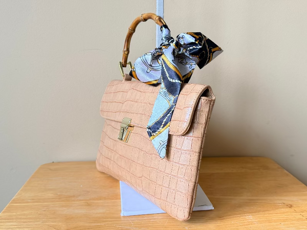Beige handbag with a bamboo handle on a wooden surface, featuring a patterned scarf draped over it.